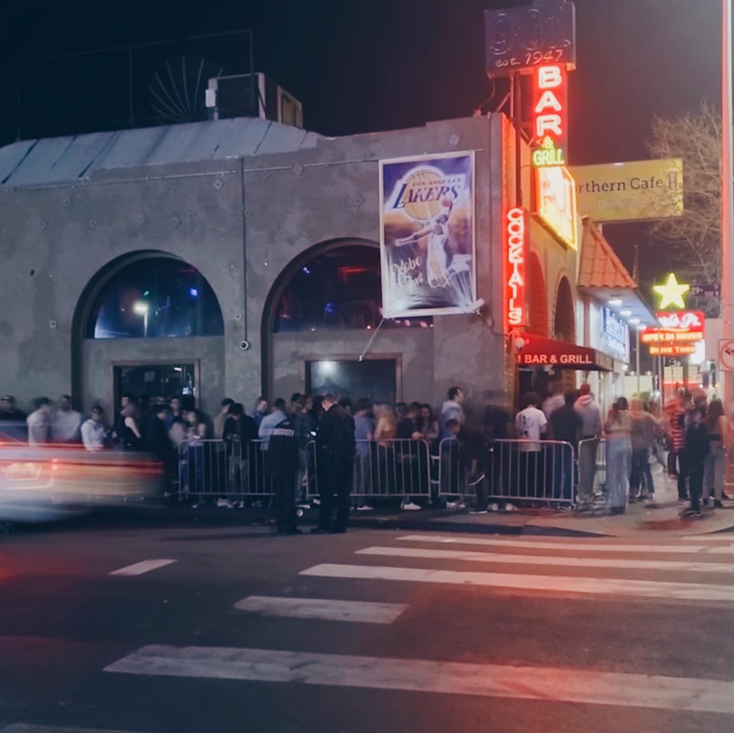 The lit 901 Bar & Grill frontage on South Figueroa at night, with a line of people waiting along the barricades outside