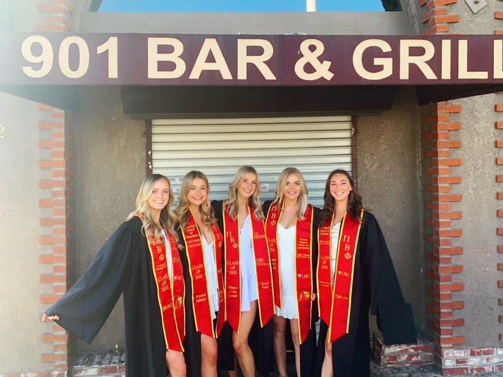 Graduates in cardinal sashes posing outside the closed roll-up gate under the 901 Bar & Grill sign